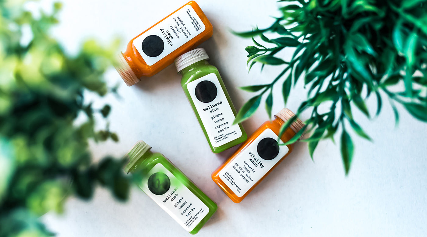 Plastic bottles of matcha and carrot juice on white background with leaves in the foreground.