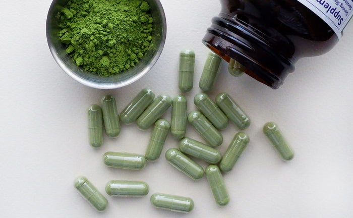 matcha pills spilling out of a bottle next to a small dish of matcha green tea on a white surface