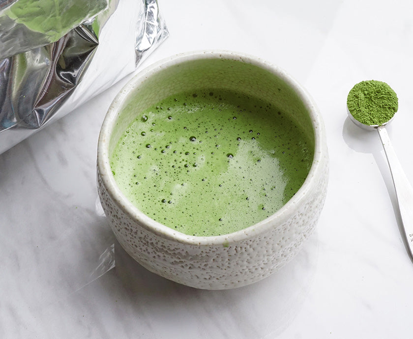 A matcha blend in a white bowl on a white table next to a spoonful of matcha and a silver bulk matcha bag